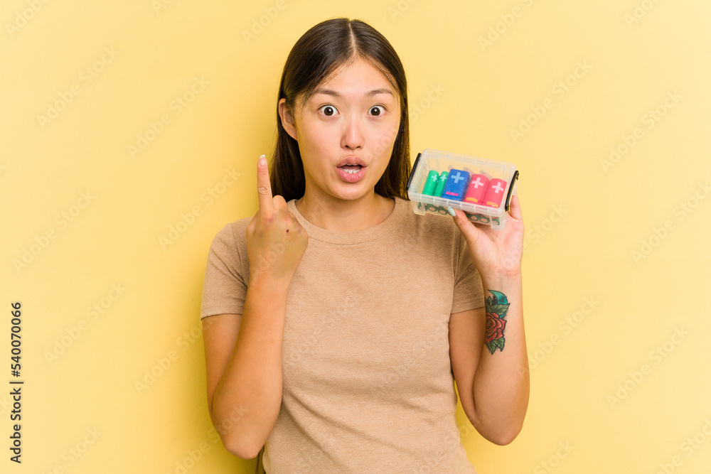 Young Asian woman holding batteries to recycle them isolated on yellow background having some great idea, concept of creativity. Young Asian woman holding batteries to recycle them isolated on yellow background having some great idea, concept of creativity.