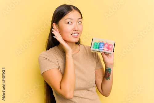 Young Asian woman holding batteries to recycle them isolated on yellow background trying to listening a gossip.