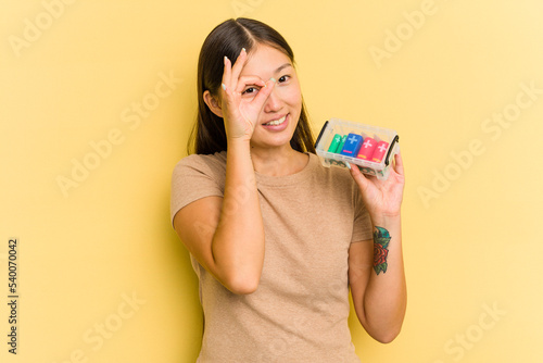 Young Asian woman holding batteries to recycle them isolated on yellow background excited keeping ok gesture on eye.