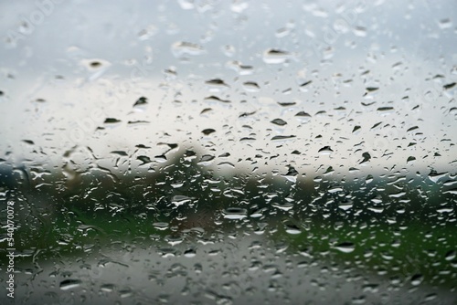 Glasscheibe mit Regentropfenmuster vor grüner Landschaft mit Feldweg zwischen grünen Ackerfeldern, Wald und Himmel bei Regen am Mittag im Herbst
