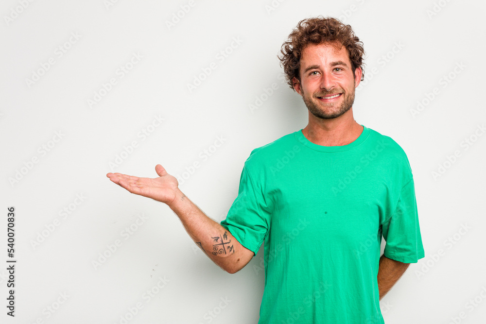 Fototapeta premium Young caucasian curly hair man isolated on white background showing a copy space on a palm and holding another hand on waist.