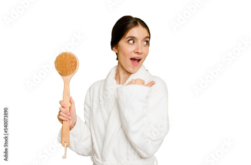 Young caucasian woman holding a shower brush isolated points with thumb finger away, laughing and carefree.