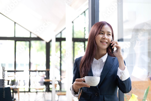 Smiling beautiful Asian businesswoman analyzing chart and graph showing changes on the market and holding smartphone at office.