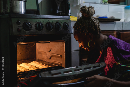 Imagen horizontal de una mujer afrocaribeña en el interior de su cocina sacando del horno unos bocadillos caribeños que acaba de preparar. 