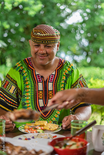 Imagen vertical de un hombre con ropa muy colorida muy sonriente degustando una deliciosa comida caribeña en su casa. 