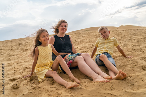 Cheerful mom, son and daughter are sitting toghether in desert sand dune.