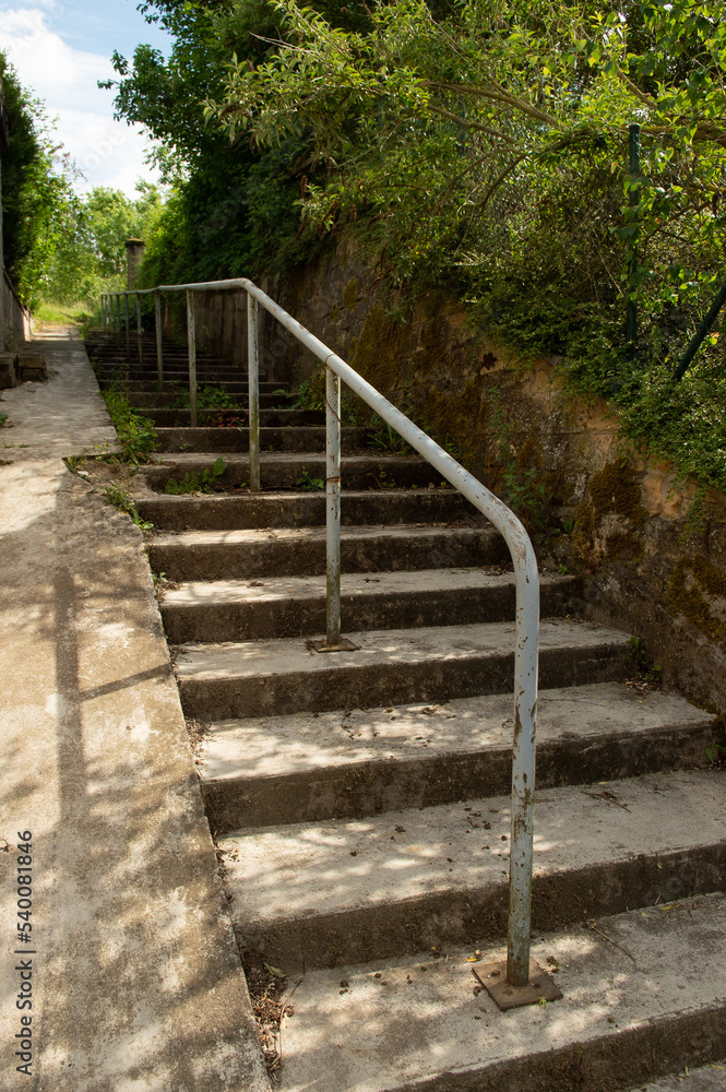 escalier extérieur en béton, avec une rampe métallique et une pente douce pour les cycles et les trotinettes ou les poussettes d'enfants; Verdure, arbres et buissons sauvages
