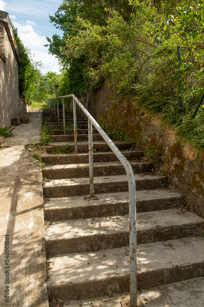 escalier extérieur en béton, avec une rampe métallique et une pente douce pour les cycles et les trotinettes ou les poussettes d'enfants; Verdure, arbres et buissons sauvages escalier extérieur en béton, avec une rampe métallique et une pente douce pour les cycles et les trotinettes ou les poussettes d'enfants; Verdure, arbres et buissons sauvages