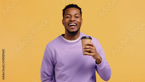 cheerful african american man in purple pullover holding paper cup with coffee to go isolated on yellow.