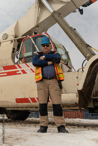 An employee in helmet and work uniform stands with his arms crossed in front of a telescopic boom crane.