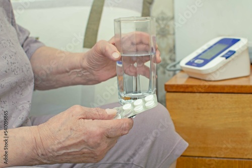 An elderly woman sitting on the bed is drinking blood pressure pills. Health problems in old age. Daily health care, taking medications. Selective focus on the drug.