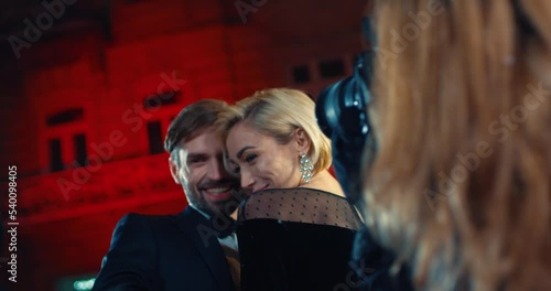 Adorable young woman and man dressed in elegant evening clothes hugging and sincerely smiling while posing for paparazzi. Celebrities posing outdoors during famous event.
