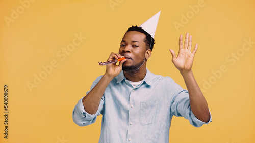 african american man in festive cap blowing party horn and gesturing isolated on yellow. photo