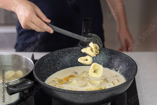 Chef cook hand making handmade tortellini ravioli with cream sauce wok pan on stove fire.