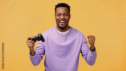 KYIV, UKRAINE - NOVEMBER 22, 2019: positive african american man holding joystick and rejoicing isolated on yellow.