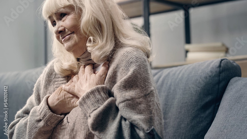 pleased senior woman in sweater holding photograph near chest at home.