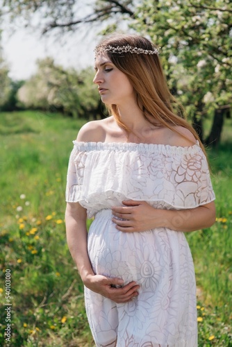 A beautiful pregnant girl in a white dress walks in an apple orchard on a sunny spring day. High quality photo