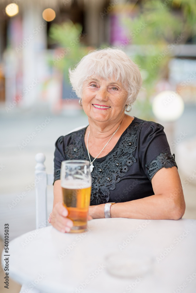 Senior woman sitting at a table in a summer cafe and drinking beer from a tall glass Senior woman sitting at a table in a summer cafe and drinking beer from a tall glass