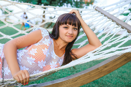 Slika na platnu A young brown-eyed brunette resting in a white hammock, which stands on the green lawn in the recreation area