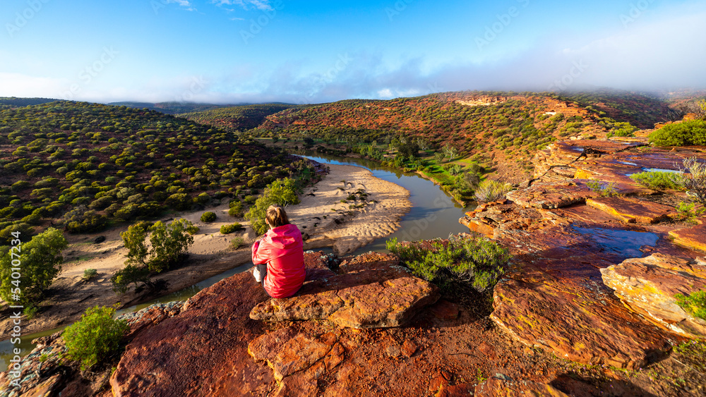 long-haired girl sits on top of a hill on the red rocks of kalbarri national park in western australia; hiking in the wilderness, australian outback