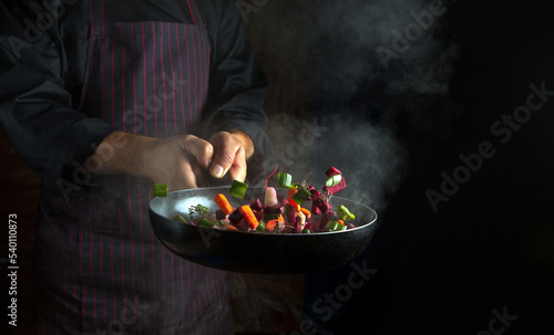 The hands of the cook throws pieces of vegetables into a hot frying pan with steam on a black background. Hotel or restaurant cooking concept