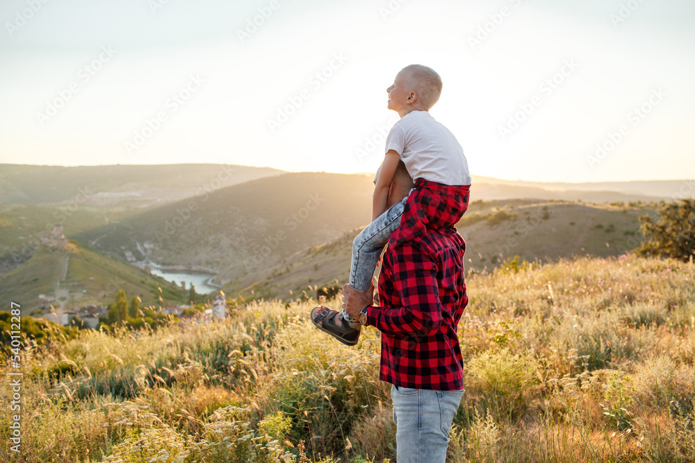 Happy family spending time together in the nature, father holding son on shoulder, have fun, playing with kid in vacation against the background the grass, mountain and sunset
