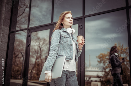 Young woman with laptop and coffee cup stands near business center