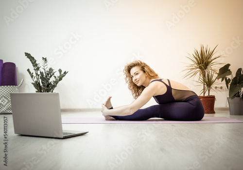 Smiling young Caucasian woman with train at home on online program on computer. Happy millennial sporty female practice yoga or pilates in living room, have webcam lesson