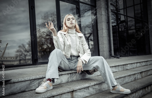 Young stylish blonde woman smokes electronic cigarette while sitting on stairs near business center