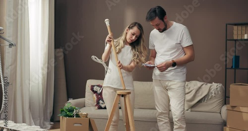 Young adorable mother with father reading instruction trying to assemble decorate furnish new floor lamp in cozy apatment carton boxes with potted plants and household stuff on background. photo