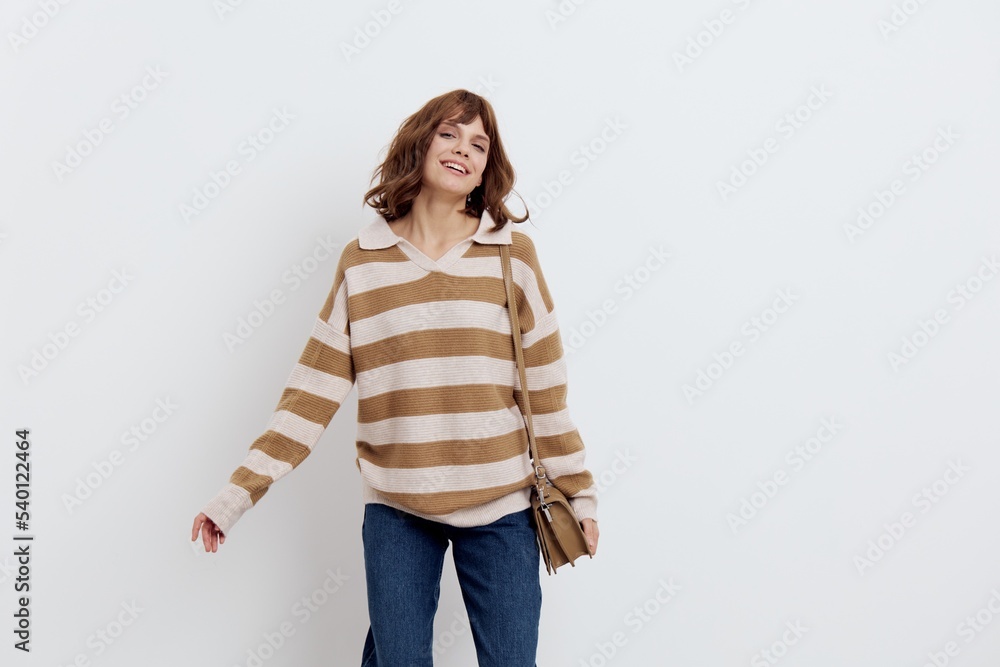a pretty sweet woman stands on a light background in a striped sweater and a bag on her shoulder, turns her head in different directions, raising her hands a pretty sweet woman stands on a light background in a striped sweater and a bag on her shoulder, turns her head in different directions, raising her hands