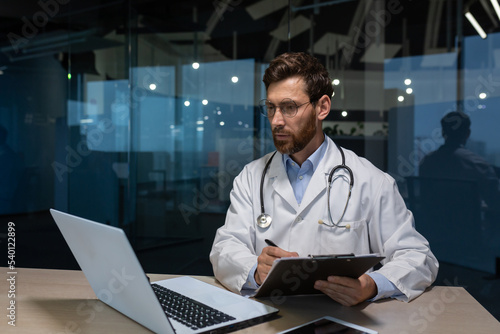 Mature doctor working on paper work inside a shabby office building, man in medical coat using laptop filling out medical documents concentrating and thinking