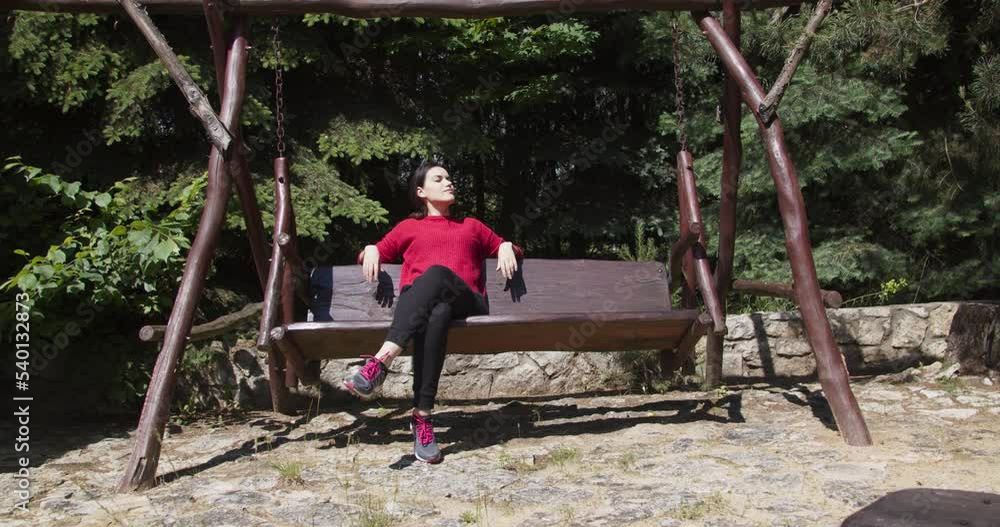 Happy woman in a red sweater sits on a wooden swing bench and enjoys a sunny day. Coniferous trees in the background.
Happy woman in a red sweater sits on a wooden swing bench and enjoys a sunny day. Coniferous trees in the background.