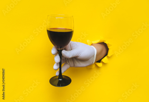 From a torn hole in bright yellow paper, a doctor\'s hand appears in a white latex glove with a glass of red wine. The concept of the benefits of alcohol, drunkenness and hangover. Copy space.