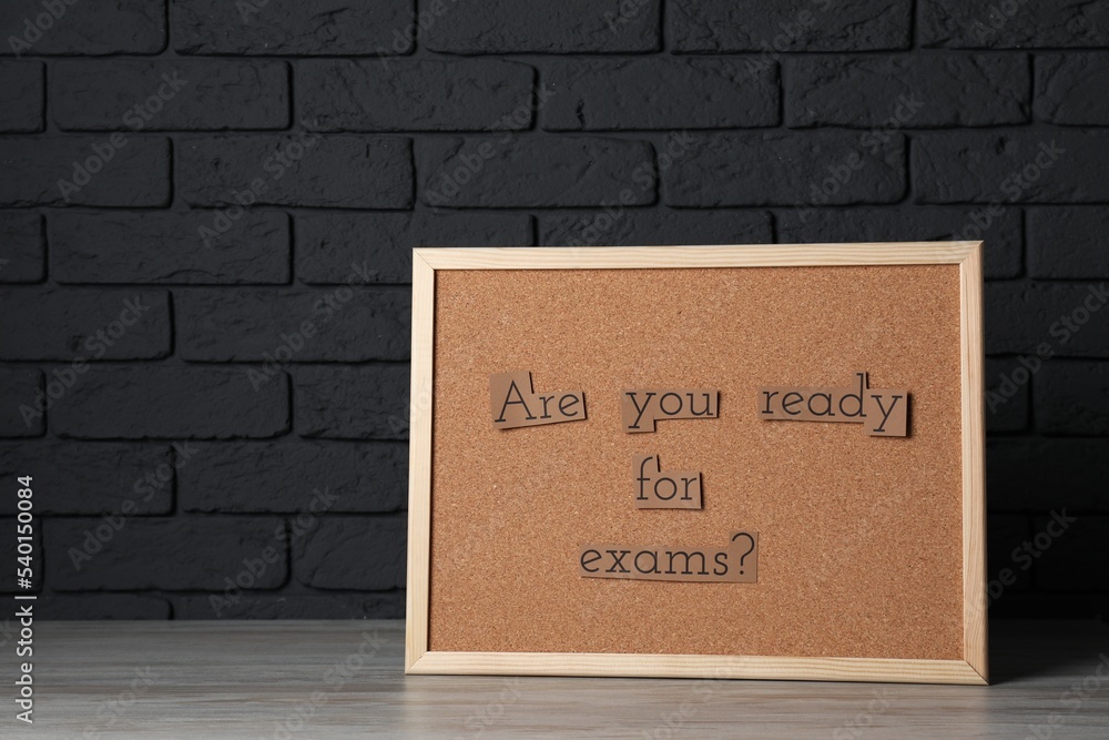 Obraz premium Cork board with phrase Are You Ready For Exams? on wooden table near black brick wall. Space for text