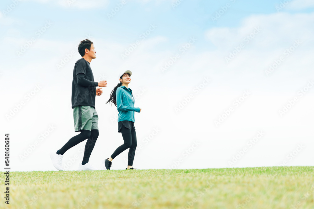 青空の見える公園でランニングするスポーツウェアを着たアジア人の男女（走る）
