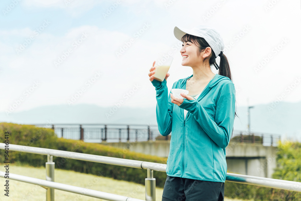 Obraz premium ボディメイクのため公園でプロテインを飲みながらトレーニングするスポーツウェアのアジア人女性 