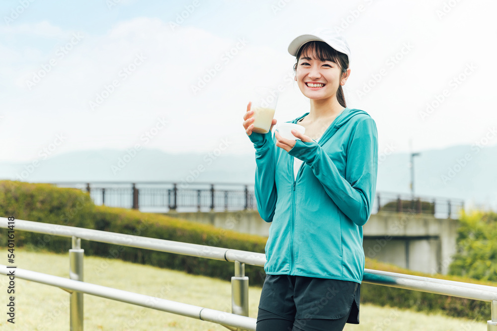 ボディメイクのため公園でプロテインを飲みながらトレーニングするスポーツウェアのアジア人女性
