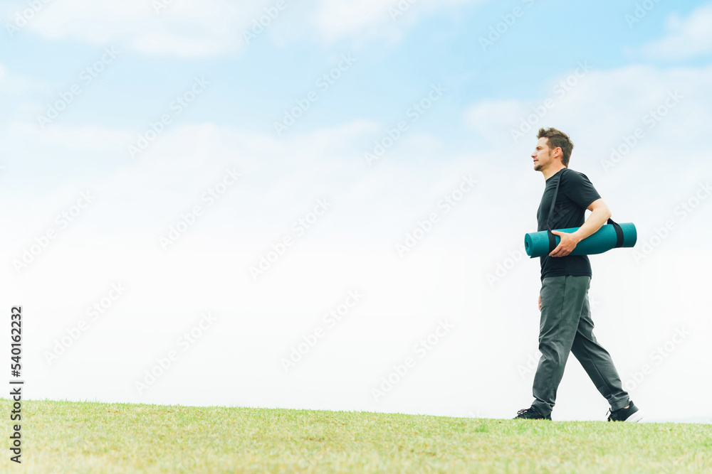 公園でヨガマットを持って歩くスポーツウェアの白人男性（パークヨガ）
