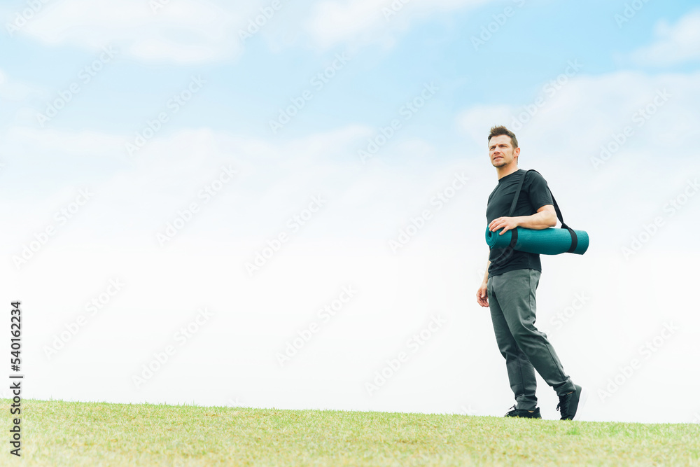 公園でヨガマットを持って歩くヨガインストラクターの白人男性

