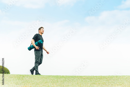 空の見える公園でヨガマットを持って歩くスポーツウェアの白人男性（笑顔） 