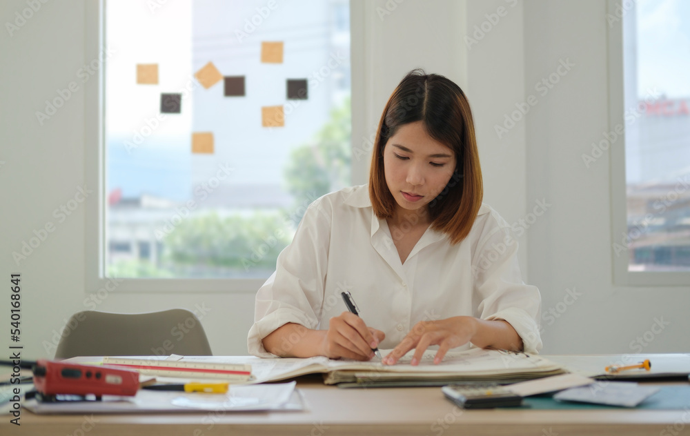 Young asian female architect sitting at desk in office and working over building designs Young asian female architect sitting at desk in office and working over building designs