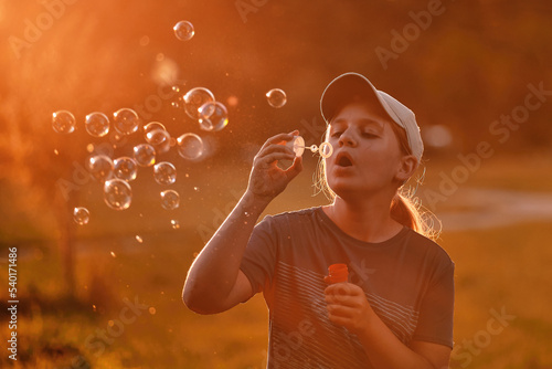 A child of 10-12 years old blows soap bubbles in the park at sunset. A school-age girl enthusiastically plays outdoors.