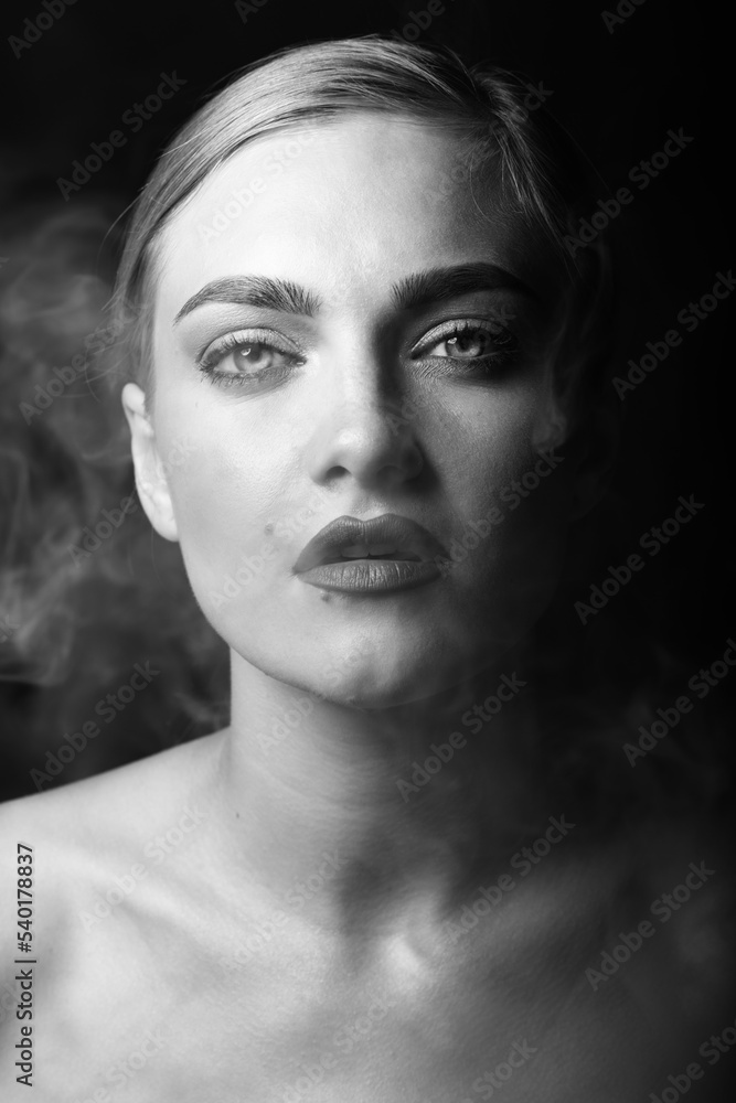 Fashion, style and make-up concept. Close-up black and white studio portrait of beautiful blonde woman blowing smoke from mouth. Model looking to camera with seductive look. Eyes in camera focus