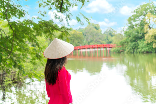 Young Asian woman tourist wearing Ao Dai (traditional Vietnamese dress) Stand by the lake, look at the Red Bridge in Hoan Kiem Lake, Hanoi, Vietnam. Copy space photo