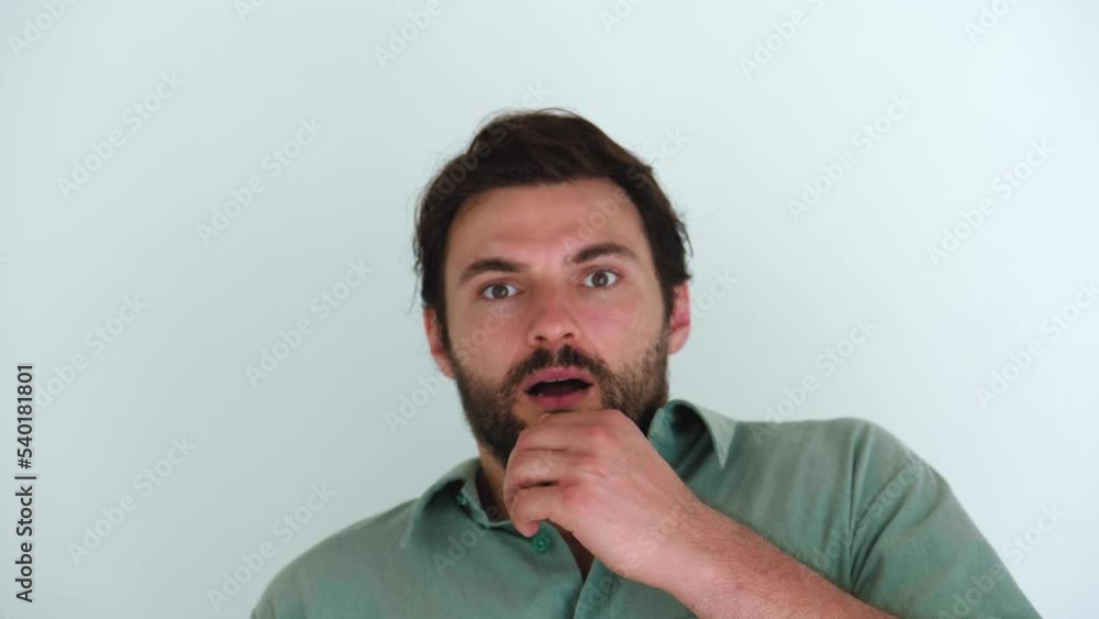 Isolated portrait over white wall background of a young dark-haired bearded Caucasian man, covering his mouth with his hand, frightened by what he saw, experiencing a sense of fear and fright. Isolated portrait over white wall background of a young dark-haired bearded Caucasian man, covering his mouth with his hand, frightened by what he saw, experiencing a sense of fear and fright.
