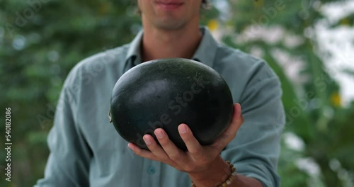 Focus on a fresh ripe organic watermelon in the hands of a handsome guy, handing it to the camera, standing against green trees backdrop. Healthy wholesome refreshing sweet dessert in hot summer days