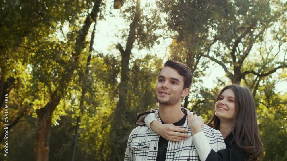 Charming caucasian lady riding on the back of her man. Joyful young couple having fun in the green park on a sunny evening. Sincere emotions and happiness concept