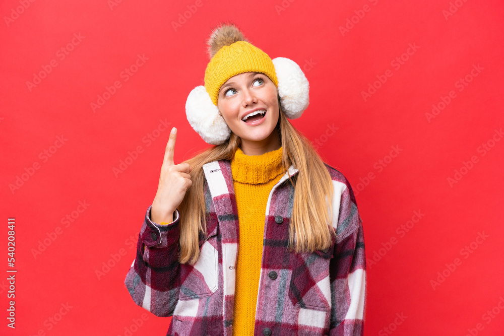 Young beautiful woman wearing winter muffs isolated on red background pointing up and surprised