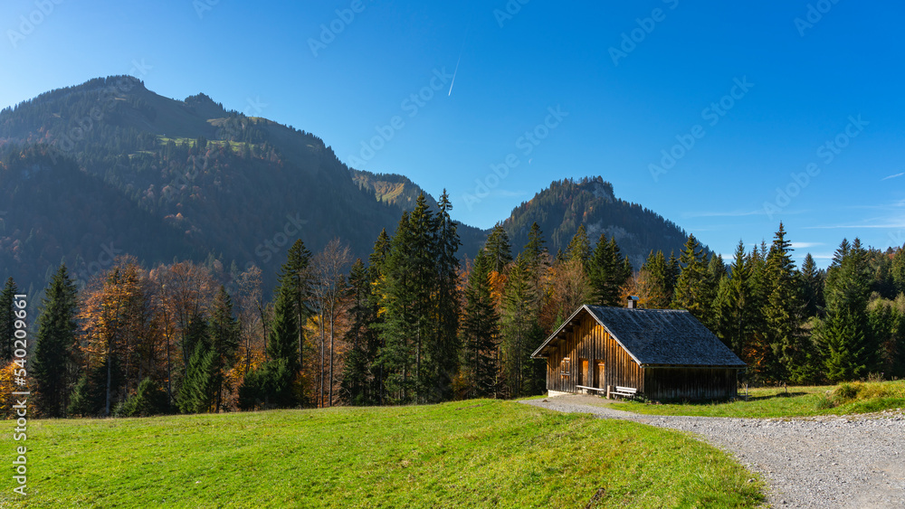 schöne hölzerne Alphütte am Weg von Sibratsgfäll nach Schönenbach. Herbst im Bregenzerwald, Österreich. Holzhaus, Stadel, Alp, am Wegrand. Berge mit herbstlich bunten Wäldern im Hintergrund